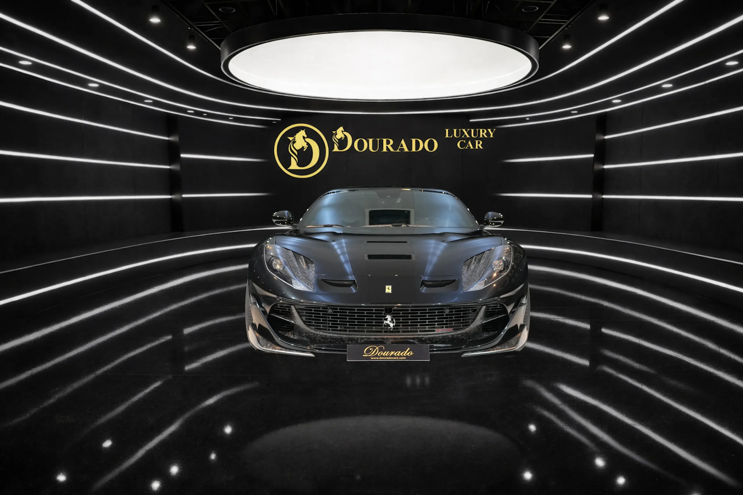 Front view of a gray luxury sports car in a black showroom with a circular ceiling light and 'Dourado Luxury Car' branding on the wall
